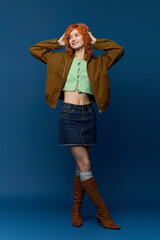 Naklejka na ściany i meble Young woman in stylish spring outfit with red hair poses gracefully against blue backdrop