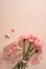 Fototapeta premium Happy Mother's Day vertical concept with pink carnations, a wrapped gift tied with satin ribbon, paper heart cutouts and soft pastel background evoking love, appreciation and gentle feminine style