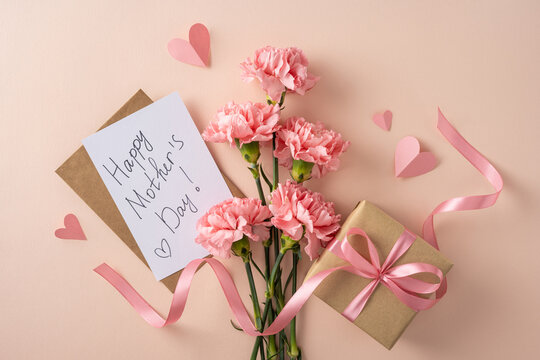 Handwritten Happy Mother's Day card with pink carnations, kraft gift box tied with ribbon and paper hearts on a soft pastel background evoking love, tenderness, celebration and feminine charm