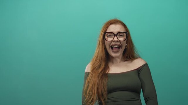 Redhead woman wearing glasses laughing broadly, bare shoulder and collarbone exposed, green off shoulder top against a teal studio backdrop; joy playfulness.