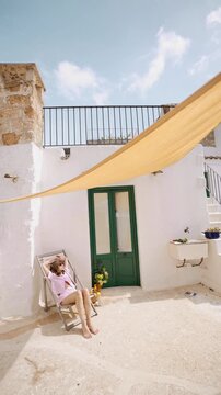 Woman relaxing on a sunlit terrace in southern Italy. Mediterranean architecture, white stone walls, soft summer light and quiet slow living atmosphere. Calm everyday life and peaceful southern