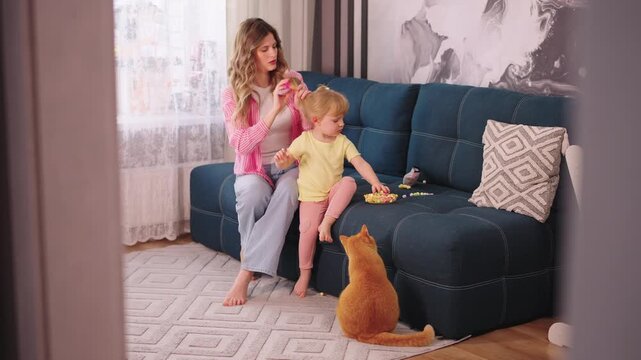 Mother and child enjoy quiet family moment on sofa. Orange cat watches them from floor beside couch. Natural light streams through window behind them. Cozy living room with soft textures