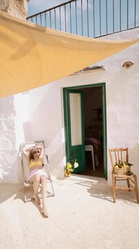 Woman relaxing on a sunlit terrace in southern Italy. Mediterranean architecture, white stone walls, soft summer light and quiet slow living atmosphere. Calm everyday life and peaceful southern