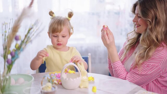 Soft light fills a cozy kitchen table. Mother and daughter share joyful Easter moments. Child laughs as she holds a small candy. Mother smiles, offering a sweet treat