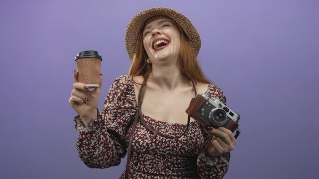 Redhead woman holding takeaway coffee cup and vintage camera in studio, smiling and laughing with straw hat and floral dress; carefree joy.