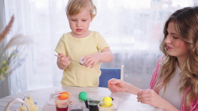 Soft light fills the room. Child holds a spoon with an egg. The mother watches with a gentle smile. They sit at a table with dye cups. The child enjoys creating Easter crafts
