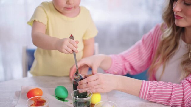 Warm light shines on a mother and child. Child stirs dye in a cup with focused curiosity. Mother watches, smiling gently. Easter egg coloring happens at home