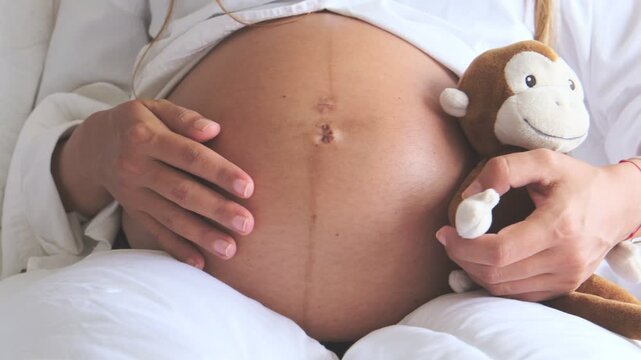 Pregnant woman gently stroking her baby bump while holding a stuffed monkey toy in bed