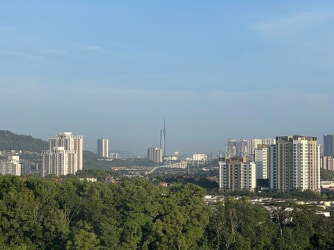 Modern residential apartment buildings standing among green trees with distant Kuala Lumpur city skyline and mountains under blue sky.