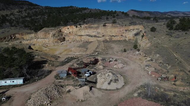 Terraced quarry face near N Foothills Hwy by Hygiene with exposed cuts, conveyors, and equipment set against steep rocky walls beside rural parcels. Pale dust, midday light in Longmont, CO