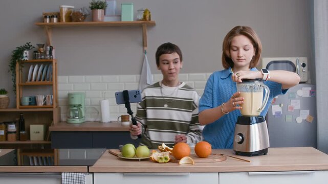 Medium shot of two Caucasian teenagers filming smoothie preparation with smartphone on stabilizer while blending fruit in kitchen, showing social media content creation and home cooking