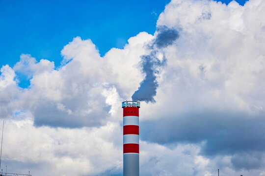 Red and white striped industrial chimney emitting dark smoke into a cloudy blue sky background