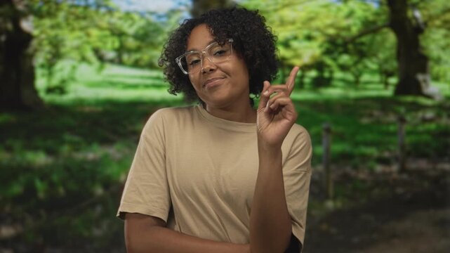 Woman wearing glasses tilts head and points finger to object near lush green trees in park; confidence insight clarity.