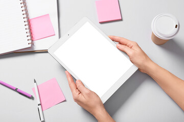 Obraz premium Female hands with tablet computer, takeaway cup of coffee, notebooks and sticky notes on grey background