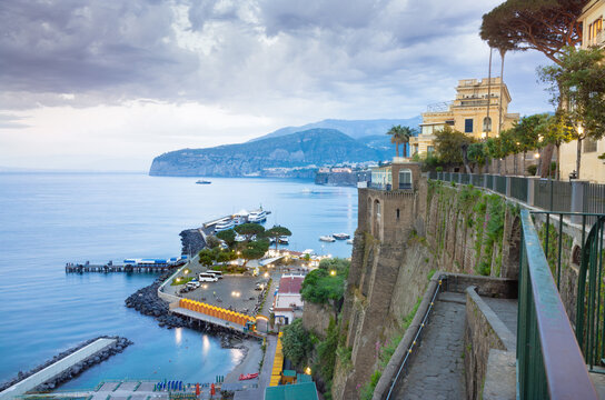 Tyrrhenian Sea and Sorrento coastline at dusk with grand villa building, winding pedestrian path, marina below, boats on bay, and distant mountains under cloudy sky 
