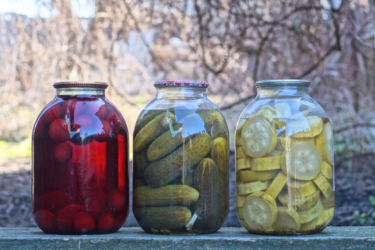 A row of three large glass jars with green pickled cucumbers, zucchini, and red compote with berries stand on a table on a daytime street