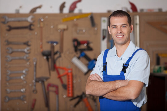 Portrait of a confident male mechanic or handyman in blue overalls standing with arms crossed in a well-equipped workshop.