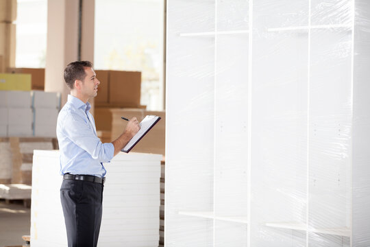 Warehouse manager or logistics supervisor writing on a clipboard while inspecting wrapped furniture or goods for shipment in a distribution center.