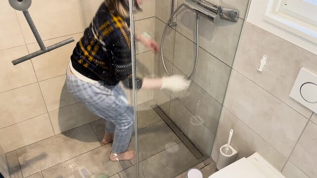 Overhead view of a woman cleaning a shower drain and scrubbing the shower floor, showing bathroom hygiene, drain maintenance, and realistic household cleaning routine in a modern home.