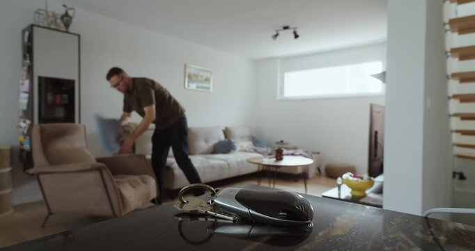 Wide shot of house keys left on a table while a man walks through the living room, then returns to pick them up, showing everyday forgetfulness, home routine, and relatable lifestyle behavior.