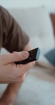Vertical close-up of a hand holding and using a remote control while relaxing at home, showing channel switching, entertainment, and everyday living room routine in a realistic lifestyle scene.