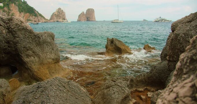 Ocean waves crash against jagged coastal rocks with view of Faraglioni peaks on horizon in Capri Italy