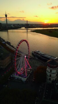 Aerial view of a ferris wheel illuminated with red and purple lights at sunset along a river with a bridge and cityscape