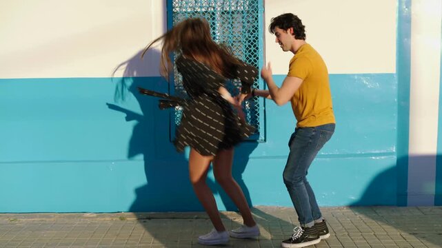 A young couple walks together on a sunny street. They laugh and play while dancing near a blue wall. The joy they share is clear as they enjoy their time outside