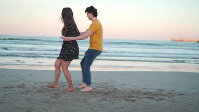 A couple of Hispanic descent dances together on the beach while the sun sets. Waves crash softly as they move joyfully along the shore, enjoying each other's company