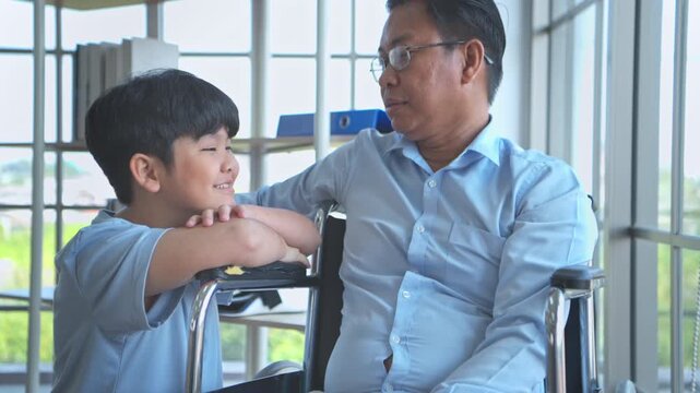 A little son speaks encouragingly to his father, who is seated in a wheelchair inside the hospital while waiting for his treatment