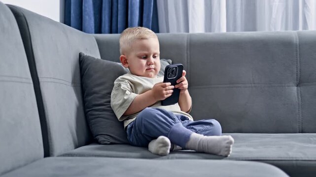 Little boy 2 years old sitting on the couch at home and playing with the phone. Toddler watching cartoons sitting on the couch. Child talking on video call on the phone. Child with phone in hands.