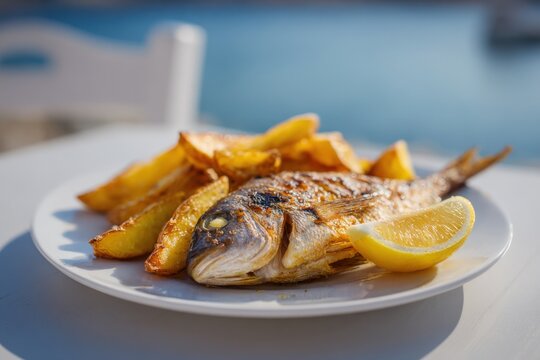 Whole grilled seabream with lemon wedges and roasted potato wedges on white plate in bright seaside setting mediterranean lunch dish