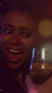 Vertical video: Woman reacting to pal raising drink, toasting at bar with lens flare lighting glass