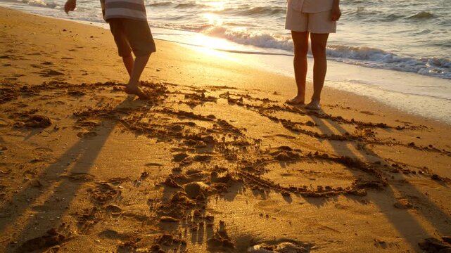 Boy and woman use their toes to draw on the beach during golden hour. This activity encourages creativity and fine motor skills in a natural outdoor setting.