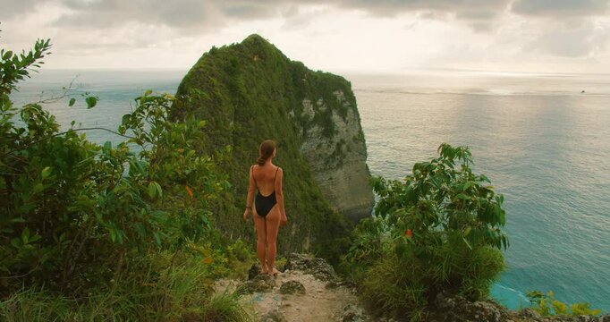 Young traveler woman stands on high cliff looking at scenic ocean bay at sunset