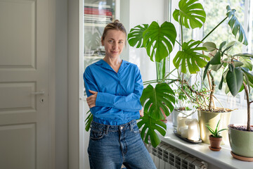 Positive middle aged woman plant lover posing beside cozy indoor garden at windowsill. Optimistic mature female green thumb smiling at camera near lush Monstera plant and various potted houseplants. © DimaBerlin