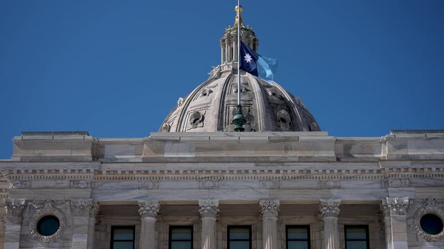 The flag of Minnesota flies on the dome of the State Capitol in Saint Paul under a bright blue sky.