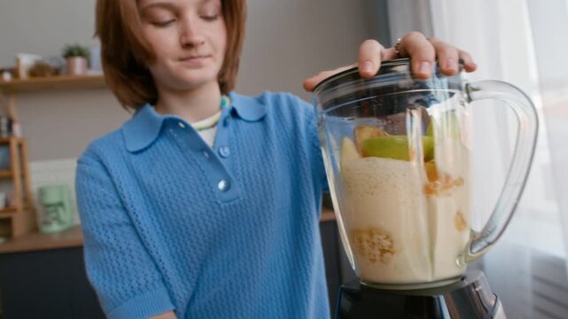 Two Caucasian teenagers making fruit smoothie in blender while filming video with smartphone stabilizer in bright kitchen, medium shot showing vlog production and home cooking