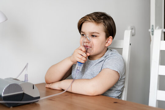 Young boy sitting at table using nebulizer for breathing treatment. Child receiving respiratory care and treatment at home. Concepts of childhood illness, respiratory treatment, home care.