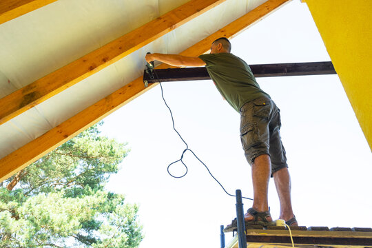 Man with cord fixes black wooden beam inside gable, close-up on hands and timber, modern yellow wall, self-build tasks.