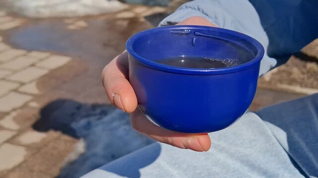 Close-up of blue thermos cup held by man during outdoor break. Concept of relaxation, tourism, travel, hiking and fresh air. Cup with hot drink on blurry background of spring thaw and urban pavement

