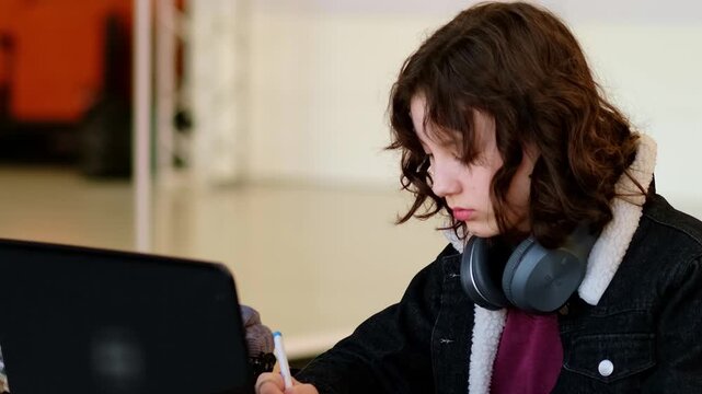Focused teenage student sitting at table with laptop and notebook, resting headphones on neck after listening to educational video, concept concentration and study motivation.