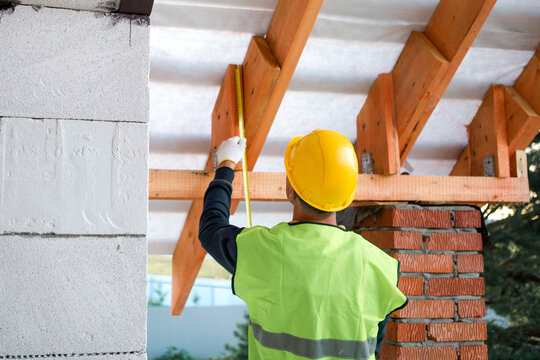 Male builder in vest checks alignment of rafters, fitting lag bolts for roof frame, visible brick support and insulation, house building.