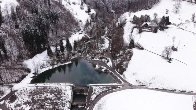 Static aerial shot of small dam at mountain base with narrow road leading into alpine landscape. Quiet natural environment and infrastructure.