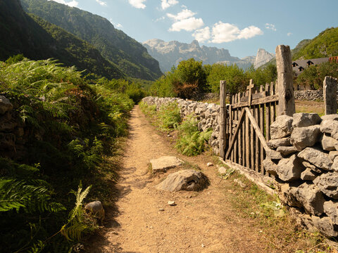 Rustic wooden gate and stone fence along a narrow dirt hiking trail in a majestic mountain valley, peaceful rural landscape with lush greenery and rocky peaks under a sunny sky