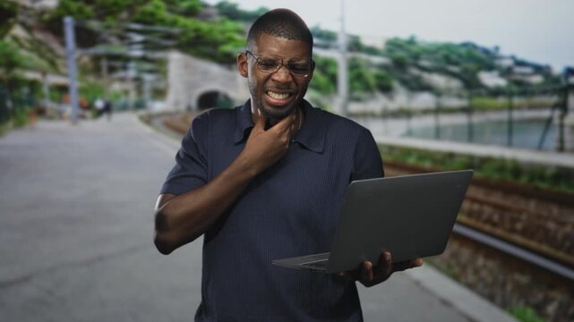 Man holding laptop clutching throat on street near train tracks and tunnel, grimacing with sore throat and pressing hand to neck; frustration urgency.