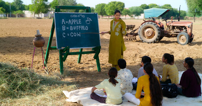 Indian young adult gen z female lady talk speak teach to villager child day under tree rural area. Happy girl boy group sitting on mat do study look board learn read english at open class field place