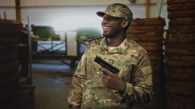Soldier man in camouflage uniform holding handgun and smiling while standing among stacked wooden crates in a building; pride duty.