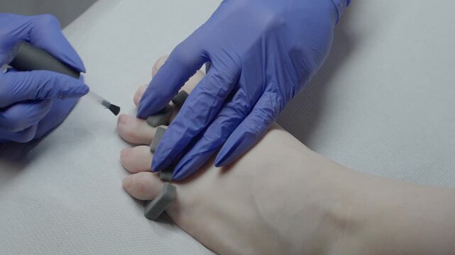 Closeup foot pedicure gloved technician. Precise polish application in sterile treatment setting. Calm clinical atmosphere, pale foot, toe separators.