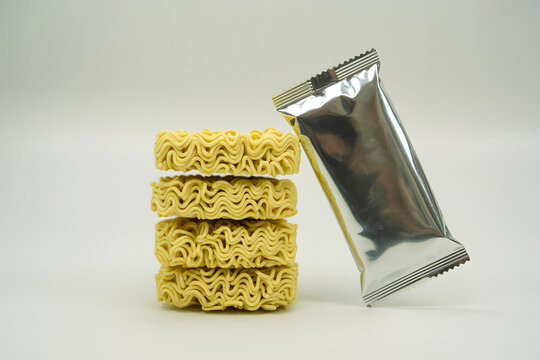A neatly stacked tower of four blocks of instant ramen noodles stands alongside a shiny, metallic silver seasoning packet, presented cleanly against a simple, bright white background.